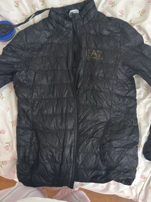 Svart pufferjacka från EA7 Armani - Snygg svart pufferjacka från EA7 Emporio Armani med guldfärgad logga på bröstet. Jackan har dragkedja framtill, hög krage och två sidofickor. Tillverkad i lätt och glansigt material som ger en modern look.de e hellt ny. 