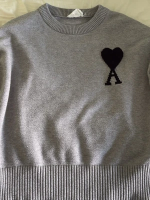 Grå stickad tröja från Ami Paris - Snygg grå stickad tröja från Ami Paris med svart hjärta och bokstaven A broderat på bröstet. Tröjan har rund halsringning, ribbade muddar och nederkant. Perfekt för dig som gillar stilrena och trendiga plagg.