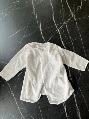 Vit omlottkofta i bomull - En enkel och mjuk vit omlottkofta med långa ärmar. Tillverkad i tunt bomullstyg och har en avslappnad passform. Perfekt för lager på lager och passar till många olika stilar.