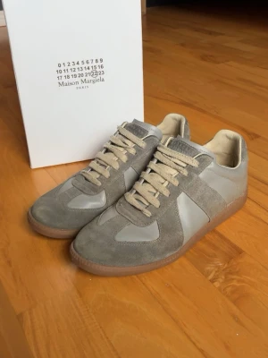 Maison Margiela grå sneakers - Maison Margiela sneakers i grå mocka och läder med beige snörning och gummisula. Klassisk low-top siluett och diskret logga på plösen. Snygg minimalistisk design som passar till det mesta. Riktigt najs nu till sommaren och nästan inget slitage  box samt dust bags följer med