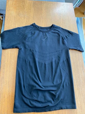 Svart tränings-tshirt från Gymshark - Snygg svart t-shirt från Gymshark med diskret mönster på bröstet och axlarna. Tillverkad i ett lätt och stretchigt material som passar perfekt till träning. T-shirten har korta ärmar och en normal passform.