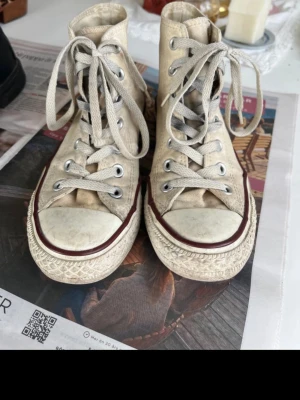 Beige Converse Chuck Taylor Highs - Klassiska beige Converse Chuck Taylor All Star high tops med vit gummisula och röda detaljer. Använda en del därav priset, men annars inga fel!