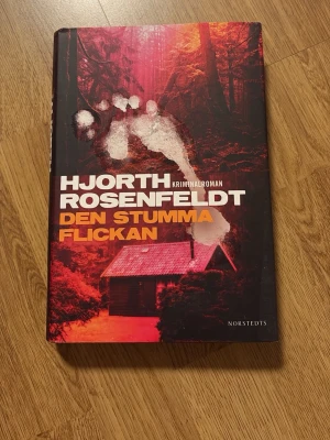 Den stumma flickan - Hjorth, Rosenfeldt - Kriminalroman av Hjorth och Rosenfeldt med hård pärm. Omslaget är mörkrött och svart med en skog, en stuga och ett vitt fotavtryck. Fjärde boken i serien. Perfekt för dig som gillar spänning och deckare.
