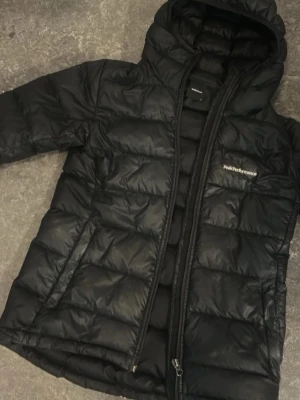 Svart dunjacka Peak Performance - Snygg svart dunjacka från Peak Performance med huva och dragkedja framtill. Jackan har quiltad design, två sidofickor och diskret logga på bröstet. Perfekt för kalla vinterdagar och riktigt varm tack vare dunfyllningen.