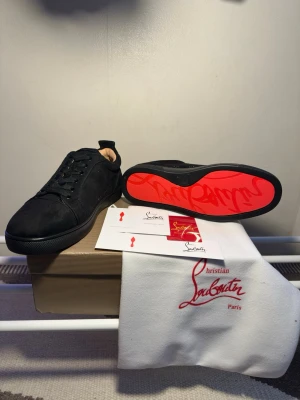 Svarta sneakers från Christian Louboutin - Stilrena svarta sneakers från Christian Louboutin med klassisk röd sula och diskret logga. Skorna har en låg profil, snörning framtill och är tillverkade i mocka. Perfekta för dig som vill ha en lyxig touch på din outfit.