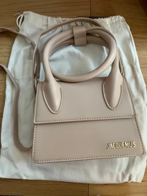 Beige Jacquemus Le Chiquito väska - Supersnygg beige handväska från Jacquemus, modellen Le Chiquito. Väskan har ett stilrent och kompakt format med guldfärgad Jacquemus-logga framtill. Tillverkad i slätt läder med både handtag och axelrem. Perfekt accessoar för att lyfta din outfit.