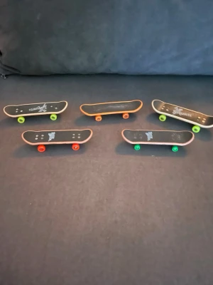 Fem fingerboards med färgglada hjul - Säljer ett set med fem fingerboards i svart med olika färger på hjulen: grönt, orange och rött. Brädorna har tryck på ovansidan och är perfekta för tricks och samling. Passar dig som gillar skate och vill öva fingerboard skills.