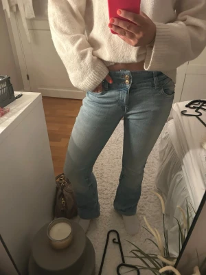 Ljusblå bootcut jeans med dubbla knappar - Säljer ett par ljusblå bootcut jeans med dubbla knappar i midjan och klassiska fem fickor. Jeansen har en lätt tvättad look och sitter snyggt på höften. Perfekta för dig som gillar en avslappnad men trendig stil.