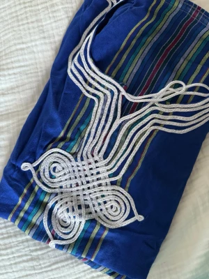 Blå klänning med broderad detalj - Unik blå marockansk  klänning med färgglada ränder och dekorativ vit broderad detalj framtill. Klänningen har ett traditionellt inspirerat mönster och är tillverkad i ett mjukt tyg som känns bekvämt mot huden. Perfekt för dig som vill sticka ut med en färgstark och personlig stil.