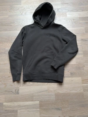 Mörkgrå hoodie från Sail Racing - Snygg mörkgrå hoodie från Sail Racing med diskret logga på bröstet. Klassisk känguruficka framtill och justerbar huva med snören. Tillverkad i mjukt bomullsmaterial som känns skönt mot huden. Perfekt för en avslappnad och stilren look.