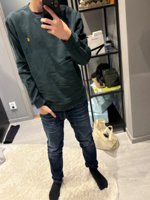 Mörkgrön sweatshirt från Lyle & Scott - Mörkgrön sweatshirt från Lyle & Scott med klassisk gul logga på bröstet. Tröjan har rund halsringning, långa ärmar och ribbade muddar vid ärmslut och nederkant. Perfekt för en avslappnad och stilren look.