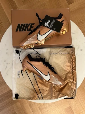 Nike Mercurial fotbollsskor - Snygga Nike Mercurial fotbollsskor i metallic guld med svart och vit swoosh. Skorna har svart snörning, rosa innersula och är tillverkade i ett lätt syntetmaterial för bästa grepp och känsla på planen. Kommer med matchande dustbag och originalkartong.