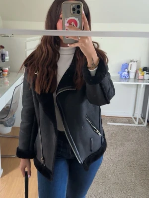 Svart skinnjacka med fuskpälsdetaljer - Snygg svart skinnjacka med fuskpäls på krage och ärmslut. Jackan har sneddragna silverfärgade dragkedjor och en cool bikerstil. Perfekt för dig som vill ha en trendig och stilren look.