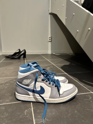 Nike Air Jordan 1 Mid blå/vit - Snygga Nike Air Jordan 1 Mid sneakers i vitt, grått och blått. Klassisk siluett med blå swoosh, blå skosnören och perforerad tåbox. Tillverkade i läder och textil med ikoniska Air Jordan-loggan på sidan. Perfekta för dig som gillar streetwear och sportig stil.