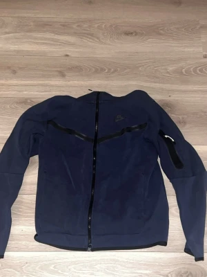 Mörkblå hoodie från Nike - Snygg mörkblå hoodie från Nike med dragkedja framtill och coola svarta detaljer över bröstet och på ärmen. Tillverkad i mjukt material som känns skönt mot huden. Perfekt för dig som gillar sportig och avslappnad stil. Huva för extra komfort.