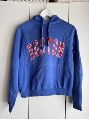 Blå hoodie med Boston tryck. S. Gina Tricot - Blå mjuk hoodie med huva och dragsnören, stor BOSTON-text i rött och vitt framtill. Klassisk college-stil med ribbade muddar och avslappnad passform. Storlek S, från Gina Tricot. Perfekt för en chill och sportig look. Lite använd. Fint skick. 