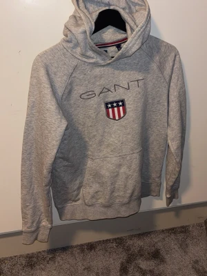 Grå hoodie från GANT med logga - Snygg grå hoodie från GANT med broderad logga och amerikanskinspirerat märke på bröstet. Klassisk känguruficka framtill och huva med dragsko. Perfekt för en avslappnad och sportig stil. Den har en liten fläck som inte går bort o tvätten! 