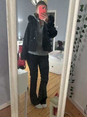 Svart kappa med fuskpäls från Gina Tricot - Stilren svart kappa med huva och mjuk fuskpäls runt kanten. Jackan har knappar vid ärmslut och framtill, samt en rak och lite boxig passform. Perfekt för dig som vill ha en snygg och mysig look under höst och vinter.