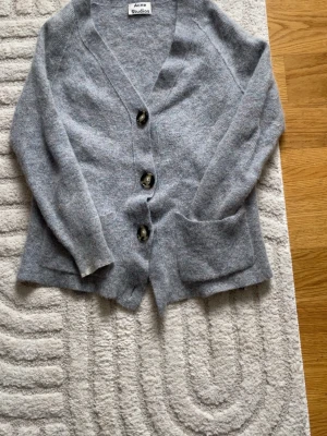 Grå kofta från Acne Studios - Mysig grå kofta från Acne Studios med stora knappar i sköldpaddsmönster och två framfickor. Koftan har en avslappnad passform, v-ringning och är stickad i mjukt material. Perfekt att slänga på sig över en t-shirt eller topp.