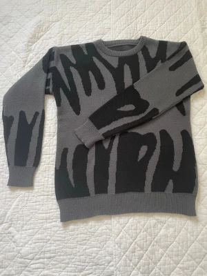 Grå stickad tröja med svart mönster - Säljer en grå stickad tröja med svart abstrakt mönster. Tröjan har rund halsringning, ribbade muddar och nederkant. Materialet ser ut att vara mjukt och bekvämt, perfekt för kyligare dagar. Passar dig som gillar en unik och modern stil.