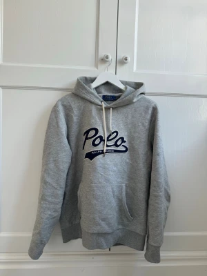 Grå hoodie från Polo Ralph Lauren - Snygg grå hoodie från Polo Ralph Lauren med stor logga i mörkblått framtill. Klassisk känguruficka och justerbar huva med vita snören. Tillverkad i mjuk bomullsblandning, perfekt för en chill och avslappnad stil.