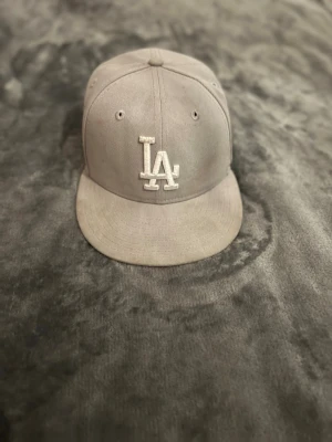 Grå LA-keps från New Era - Snygg grå keps från New Era med LA-logga broderad framtill i vitt. Klassisk modell med böjd skärm och ventilerande hål. Passar perfekt till streetwear och har en stilren look. Tillverkad i slitstarkt tyg.