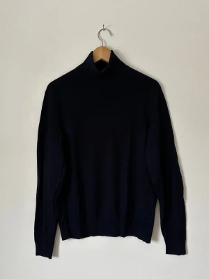 Mörkblå polotröja i stickat material - Klassisk mörkblå Uniqlo stickad polotröja med lång ärm och hög krage. Tröjan har en enkel och stilren design, perfekt för lager på lager och passar till det mesta. Materialet ser mjukt och följsamt ut, vilket gör den skön att bära under kyligare dagar. 8B19