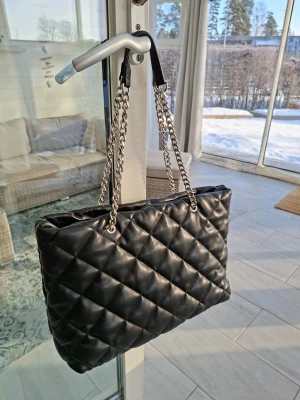 Svart quiltad handväska med kedjerem - Snygg svart handväska i quiltat konstläder med silverfärgade kedjeremmar och svarta axelband. Väskan har en klassisk rektangulär form och ger en lyxig känsla. Perfekt för dig som vill ha en trendig och rymlig väska till alla dina grejer.