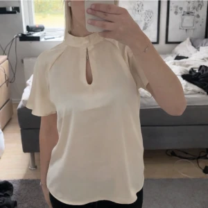 champagnefärgad blus - Stilren champagnefärgad blus i glansigt material med korta ärmar och hög krage. Framtill finns en elegant nyckelhålsöppning. Perfekt att matcha med både jeans och kjol för en trendig look.