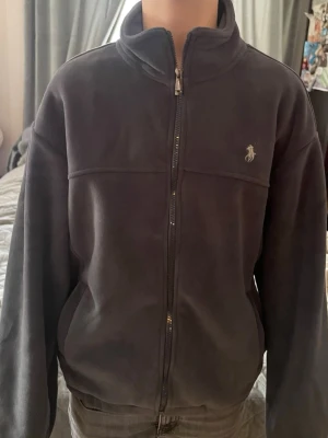 Grå fleecejacka från Polo Ralph Lauren - Mysig mörkblå fleecejacka från Polo Ralph Lauren med dragkedja framtill och hög krage. Jackan har en beige insida och den klassiska Polo-loggan broderad på bröstet. Perfekt för kyliga dagar och enkel att matcha med olika outfits. Strl M