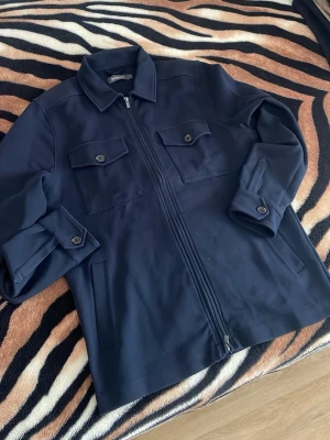 Mörkblå overshirt från Dressmann - Stilren mörkblå overshirt från Dressmann med dragkedja framtill, två bröstfickor med lock och knapp samt sidofickor. Klassisk krage och knappdetaljer vid ärmsluten. Perfekt för lager-på-lager och enkel att matcha till olika outfits.