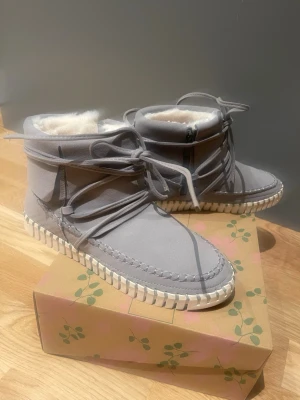 Gråa fodrade stövletter med snörning stl 38 oanvända  - Säljer ett par gråa stövletter med snörning och vit sula. Insidan är fodrad med mjukt, vitt fuskpälsfoder som håller dig varm. Skorna har en modern design med dekorativa sömmar och dragkedja på sidan för enkel påtagning. Perfekta för kalla dagar.