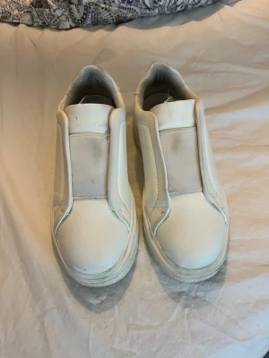 Vita slip-on sneakers utan snörning - Stilrena vita slip-on sneakers med elastiskt band över vristen och tjock sula. Skorna har en minimalistisk design utan snörning och är tillverkade i syntetmaterial med textildetaljer. Perfekta för en clean och enkel look. Stor så mig som vanligtvis har 39 men passar en med storlek 40.