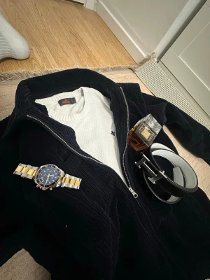 Vit stickad tröja från Morris - Säljer en stilren vit stickad tröja från Morris med ett diskret broderat emblem på bröstet. Tröjan har långa ärmar och ribbade muddar vid ärmslut och nederkant. Perfekt för en klassisk look.