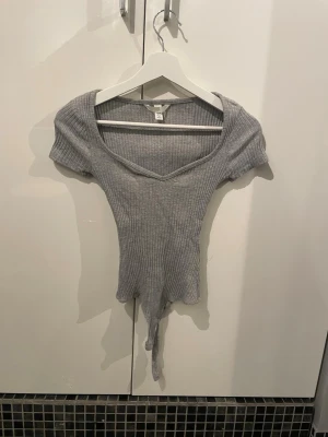 Grå ribbad body med v-ringning - Säljer en grå ribbad body med korta ärmar och djup v-ringning. Bodyn har en åtsittande passform och knäppning i grenen. Perfekt att styla till jeans eller kjol för en trendig look.