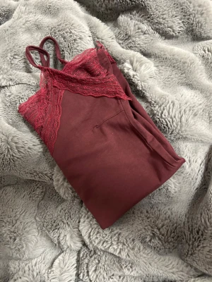 Vinrött nattlinne med spetsdetaljer - Säljer ett vinrött nattlinne med tunna axelband och fina spetsdetaljer vid urringningen. Materialet känns mjukt och bekvämt, perfekt för mysiga kvällar hemma. Spetsen ger en extra touch av elegans och nattlinnet har en enkel, rak passform.