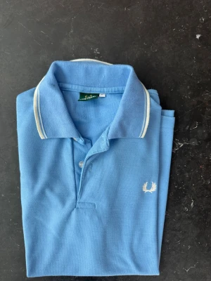 Blå piké från Fred Perry - Snygg blå pikétröja från Fred Perry med vit logga broderad på bröstet och vita detaljer på kragen och ärmsluten. Klassisk pikékrage och korta ärmar. Perfekt till en avslappnad men ändå stilren look.