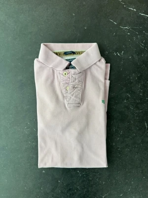 Ljusrosa pikétröja från Abercrombie - Säljer en ljusrosa pikétröja från Abercrombie & Fitch med klassisk krage, korta ärmar och knappar framtill. Tröjan har en liten grön broderad logga på bröstet och är gjord i mjuk bomull. Perfekt för en avslappnad och stilren look.