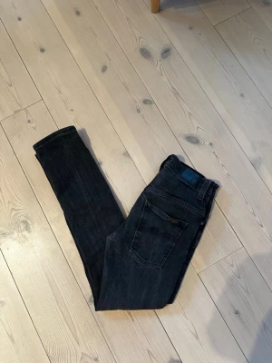 Svarta jeans från Nudie Jeans - Snygga svarta jeans från Nudie Jeans med klassisk femficksdesign och fin passform. Jeansen har en mörk tvätt och är tillverkade i stretchig bomullsdenim för extra komfort. Perfekta för dig som gillar en stilren och modern look. OBS, jeansen är små i storleken 