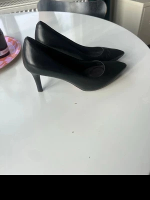 Svarta klassiska pumps med smal klack - Säljer ett par svarta pumps med spetsig tå och smal klack. Skorna har en stilren design och är tillverkade i ett slätt material som liknar skinn. Insidan har en trutech sula som ger stöd och gör skorna bekväma. Perfekta för dig som gillar tidlös och elegant stil.