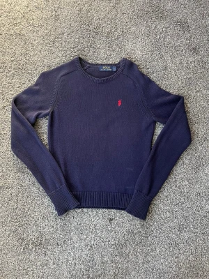 Mörkblå stickad tröja Polo Ralph Lauren - Snygg mörkblå stickad tröja från Polo Ralph Lauren med rund halsringning och det klassiska röda broderade logot på bröstet. Tröjan har långa ärmar och ribbade muddar vid ärmslut. Storleken är Dam S