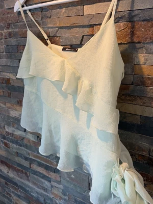 Ljusgrön volangtop från Gina Tricot - Hej!☀️Jag säljer en ljusgrön topp från Gina Tricot med tunna axelband och volangdetaljer framtill och nedtill. Toppen har en asymmetrisk nederkant och en stor rosett på sidan. Endast använd vid två tillfällen💕superfin!!