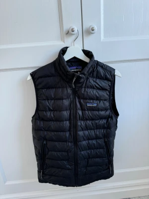 Svart dunväst från Patagonia - Säljer en svart quiltad dunväst från Patagonia med dragkedja framtill och två sidofickor med dragkedja. Västen är ärmlös och har en hög krage. Perfekt för lager-på-lager och outdoor vibes.