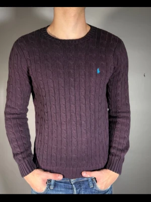 Lila kabelstickad tröja från Ralph Lauren - Snygg lila kabelstickad tröja från Ralph Lauren med rund halsringning och blå broderad logga på bröstet. Tröjan har långa ärmar och ribbade muddar vid ärmslut och nederkant. Perfekt för dig som gillar klassisk och stilren look.