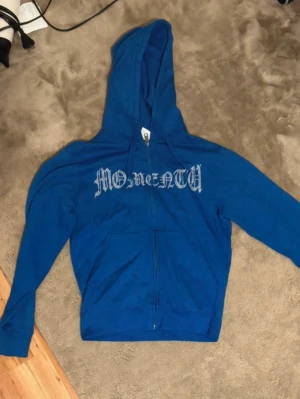 Blå rhinestone hoodie - Blå hoodie med dragkedja från Russell i storlek XS. Framsidan har trycket 'MOMENTUM' i gotisk stil. Hoodien har huva och två fickor framtill. Mjuk insida för extra komfort, perfekt för en avslappnad streetwear-look.