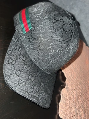 Svart Gucci keps  - Snygg svart keps från Gucci med klassiskt GG-monogram över hela kepsen och gröna samt röda ränder på sidan. Tillverkad i bomull och polyester med elastan för extra komfort. Perfekt accessoar för dig som vill ha en exklusiv och trendig look.