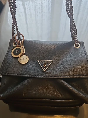 Svart handväska från Guess - Snygg svart handväska från Guess med kedjerem och dekorativa metallbrickor. Väskan har ett triangelformat Guess-emblem framtill och flera fack inuti, inklusive ett med dragkedja. Tillverkad i konstläder med ljusbeige foder och logotypdetaljer.