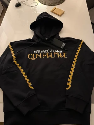 Svart Versace Jeans Couture hoodie - Snygg svart hoodie från Versace Jeans Couture med guldfärgade kedjedetaljer längs ärmarna och stort logotryck i vitt och guld på bröstet. Klassisk huva med dragsko och ribbade muddar. Perfekt för dig som vill sticka ut med exklusiv streetwear. Större i storlek helt ny oanvänd äkta.