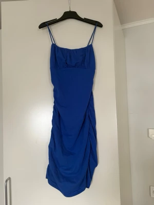 Blå kort klänning med smala axelband - Snygg blå kort klänning med smala axelband och draperad detalj på sidan. Klänningen har en enkel, figurnära passform och är perfekt för dig som vill ha en stilren men ändå trendig look. Materialet känns mjukt och följsamt.