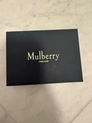 Svart läderarmband från Mulberry - Stilrent svart armband i läder från Mulberry med ikoniskt guldfärgat metallspänne och Mulberry-logga. Armbandet har en bred design och känns både lyxigt och tidlöst. Perfekt accessoar för dig som gillar klassiska detaljer och exklusiva material. Strl S